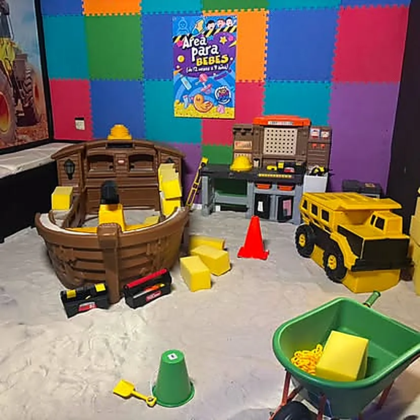 Área para bebés: arenero con un barco pirata de juguete, mesa de herramientas, camión escolar amarillo y el letrero Área para Bebés en la pared de gomaespuma de colores.