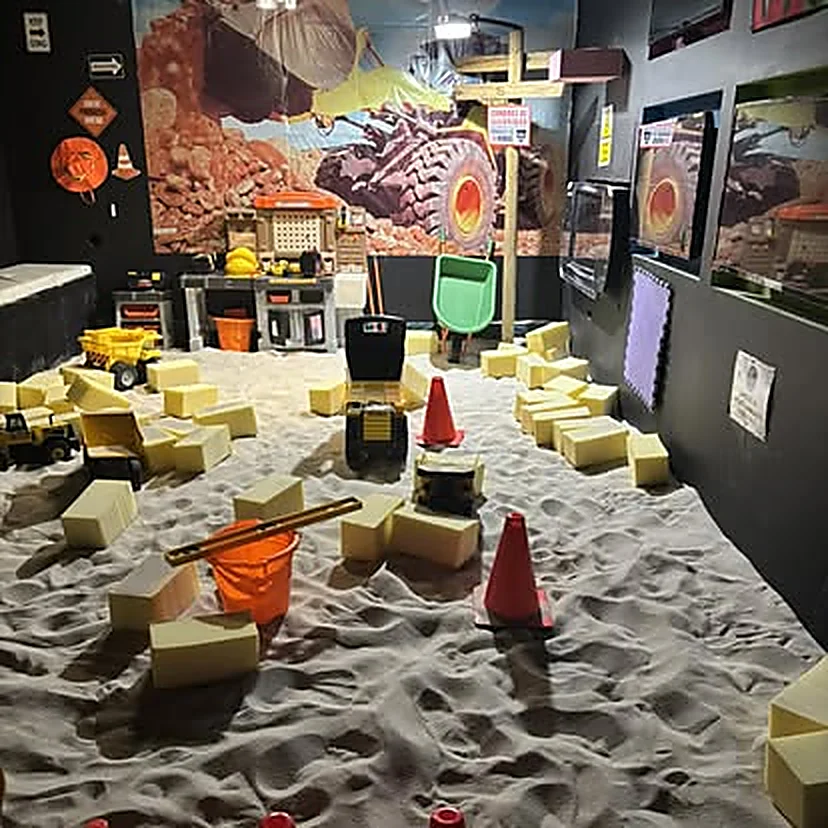 Zona de construcción: arena, bloques amarillos de espuma, camiones de juguete, conos y un mural de camión minero.
