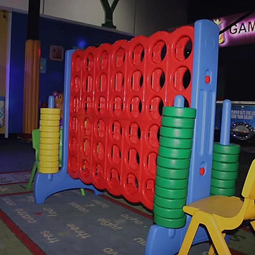 Conecta 4 gigante de piso en rojo, azul, amarillo y verde, con una silla infantil al lado y señalización luminosa al fondo.
