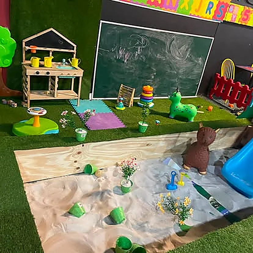 Rincón de juego de rol: cocinita de madera con utensilios, pizarra verde, animal de peluche, aros de colores y regadera de juguete sobre pasto sintético.
