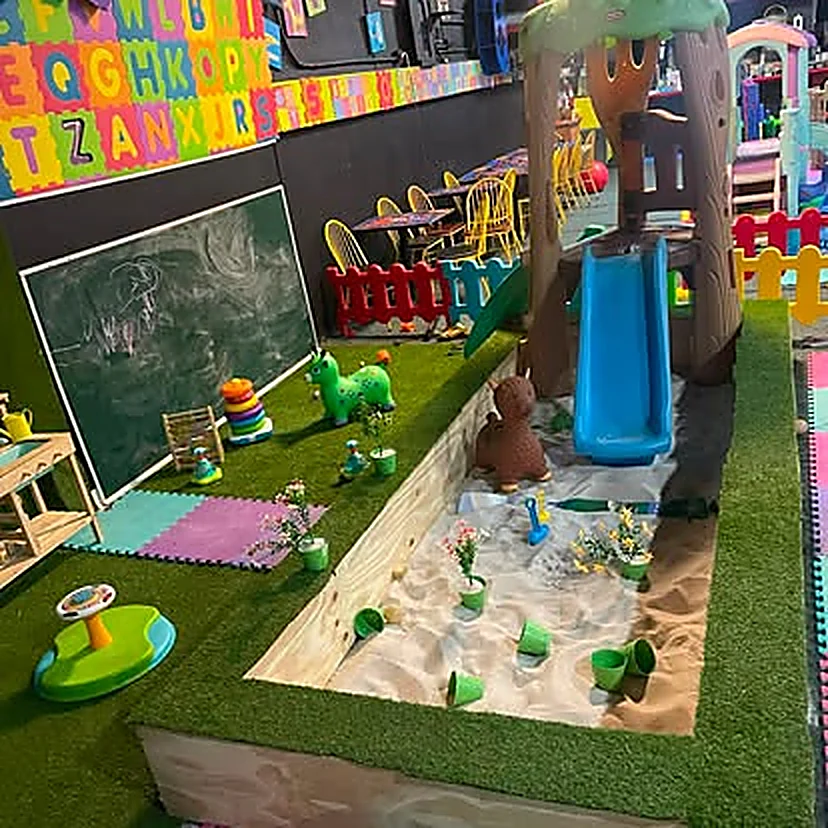 Área para pequeños: pasto sintético, casita de árbol con tobogán azul, pizarra de tiza y un arenero con flores.